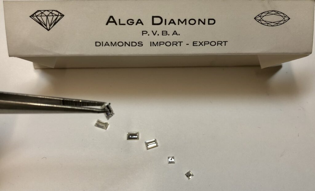 Een diamant van emerald-vorm wordt met een pinset vastgenomen. Op de tafel staan nog drie diamanten van emerald vorm en twee diamanten van prinses vorm. Op de achtegrond is de naam en logo van het bedijf, Alga Diamond, zichtbaar.